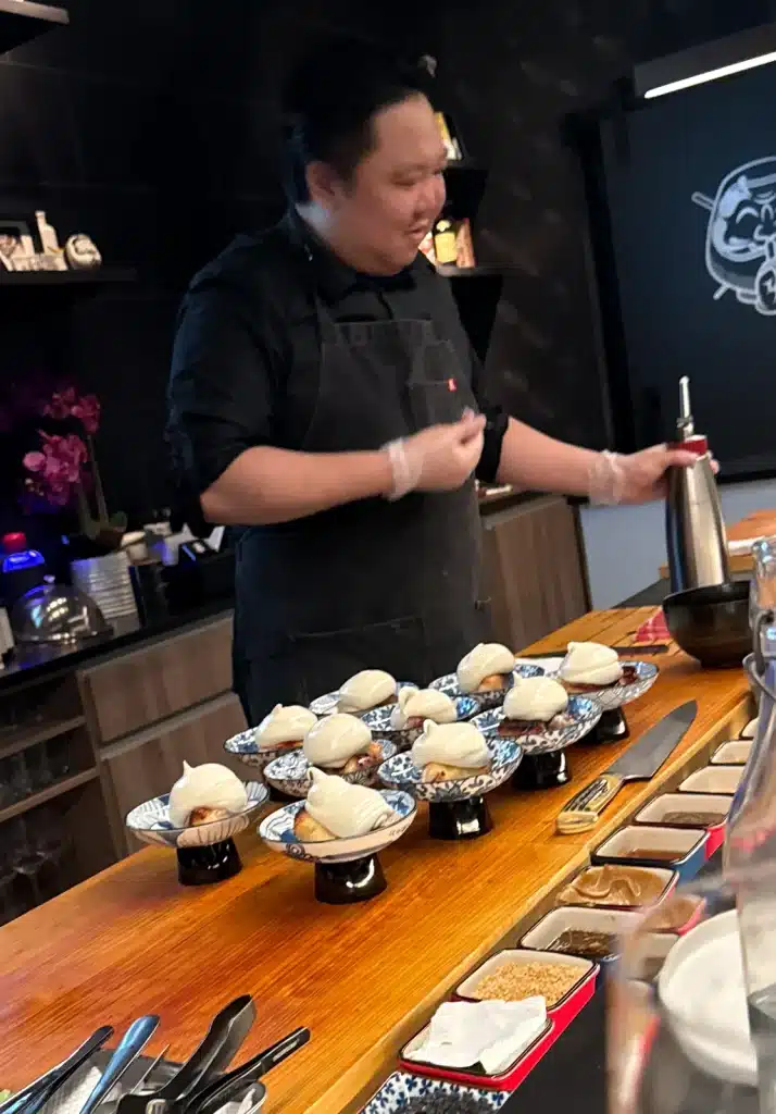  A chef stands behind a row of patterned bowls topped with pale foam, holding a siphon.
