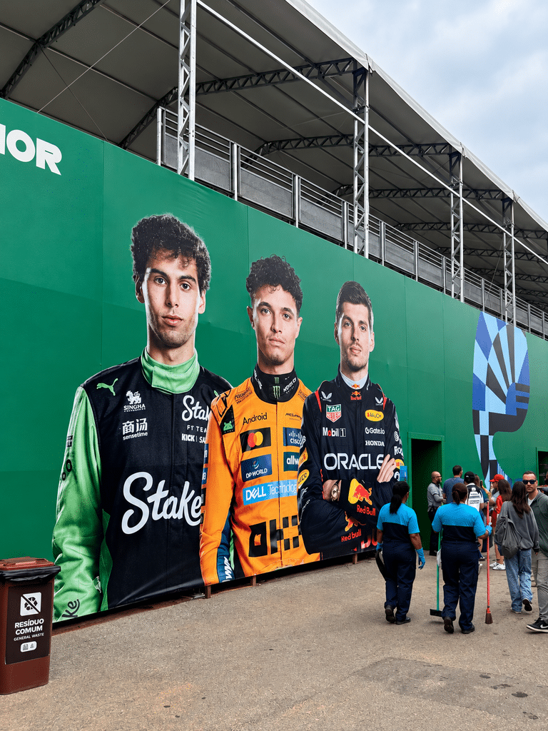 Large Formula 1 driver portraits on a green wall beneath a covered grandstand at Interlagos, with visitors walking past.