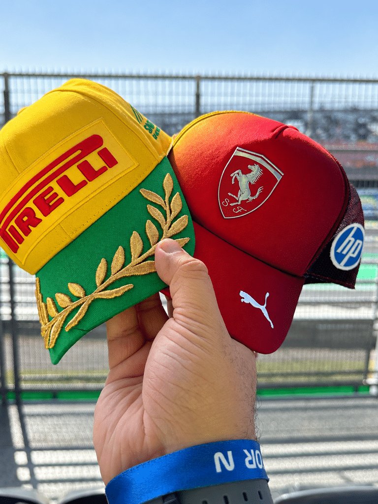 A hand holding a yellow and green Pirelli cap with gold laurel embroidery beside a burgundy Ferrari cap at Interlagos.