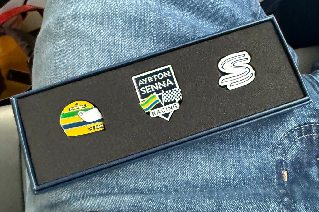 Three Ayrton Senna pins in a dark presentation box resting on blue jeans, including a yellow helmet, racing crest and stylized S.