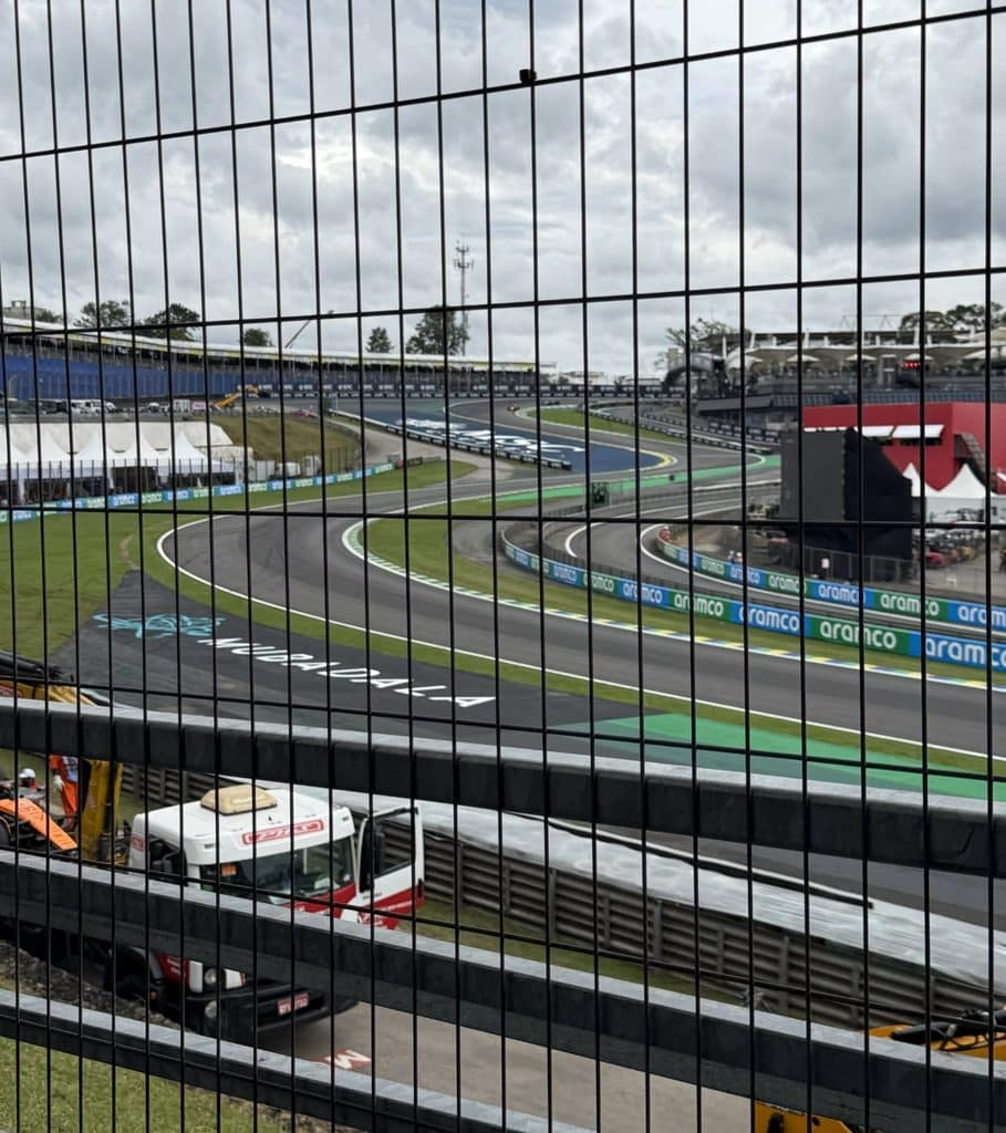 Formula 1 Grandstand The Senna S at Interlagos seen through a tall black safety fence, with a service truck below and cloudy skies overhead.